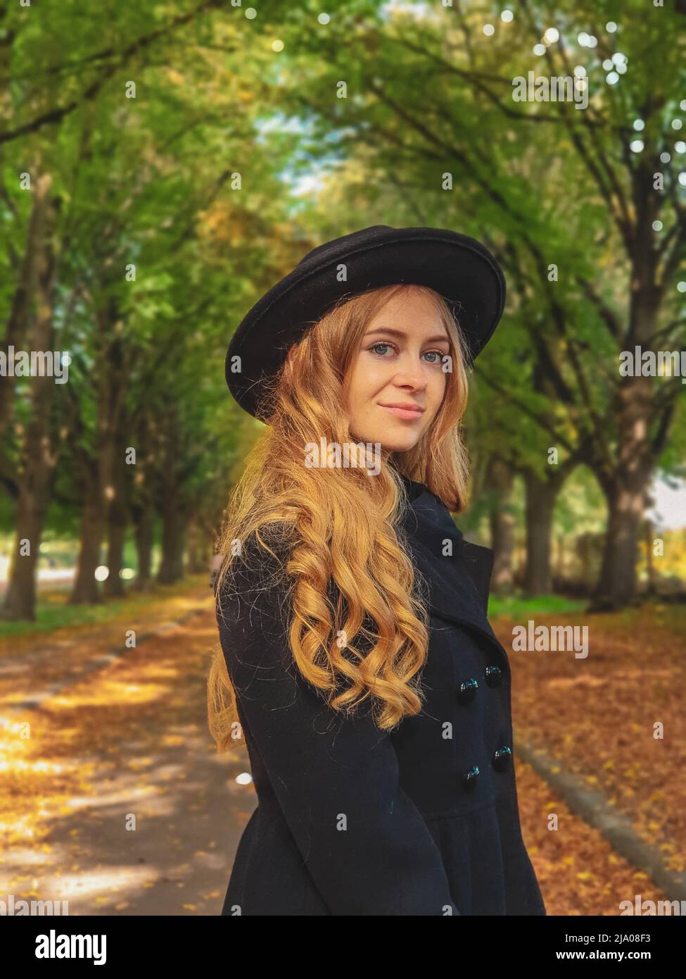 Mujer joven con hojas de otoño en el fondo. Hermosa chica en chaqueta negra con pelo rizado y otoño verde amarillo arce parque fondo Foto de stock