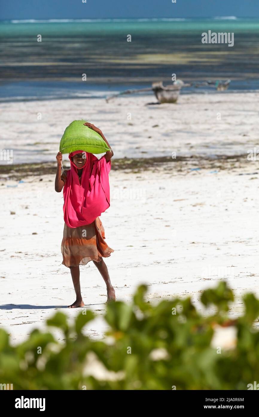 Una chica que llevaba un saco de algas en la cabeza en la playa de Jambiani con un barco tradicional 'Ngalawa' al fondo, Zanzíbar, Tanzania, África. Foto de stock