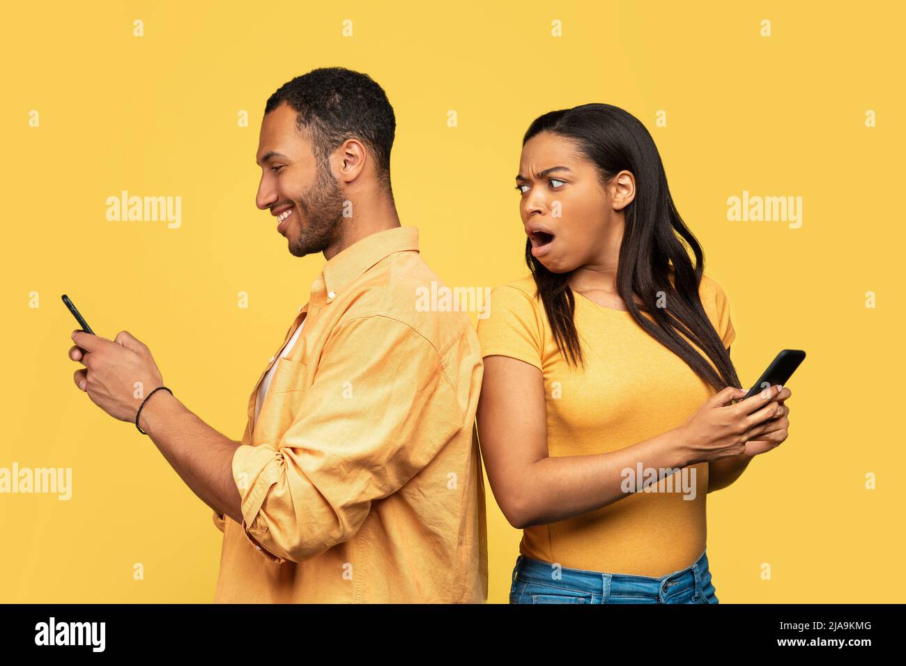 Una mujer negra conmocionada espiando a su novio que está usando un smartphone, charlando con un amante, engañando sobre fondo amarillo Foto de stock