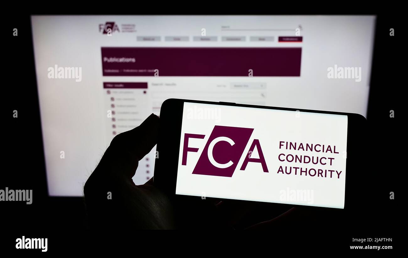 Persona que sostiene el teléfono móvil con el logotipo de British Financial Conduct Authority (FCA) en la pantalla delante de la página web. Enfoque en la pantalla del teléfono. Foto de stock