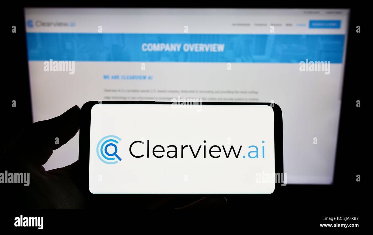 Persona que sostiene un smartphone con el logotipo de la empresa de reconocimiento facial Clearview AI Inc. En la pantalla frente al sitio web. Enfoque en la pantalla del teléfono. Foto de stock