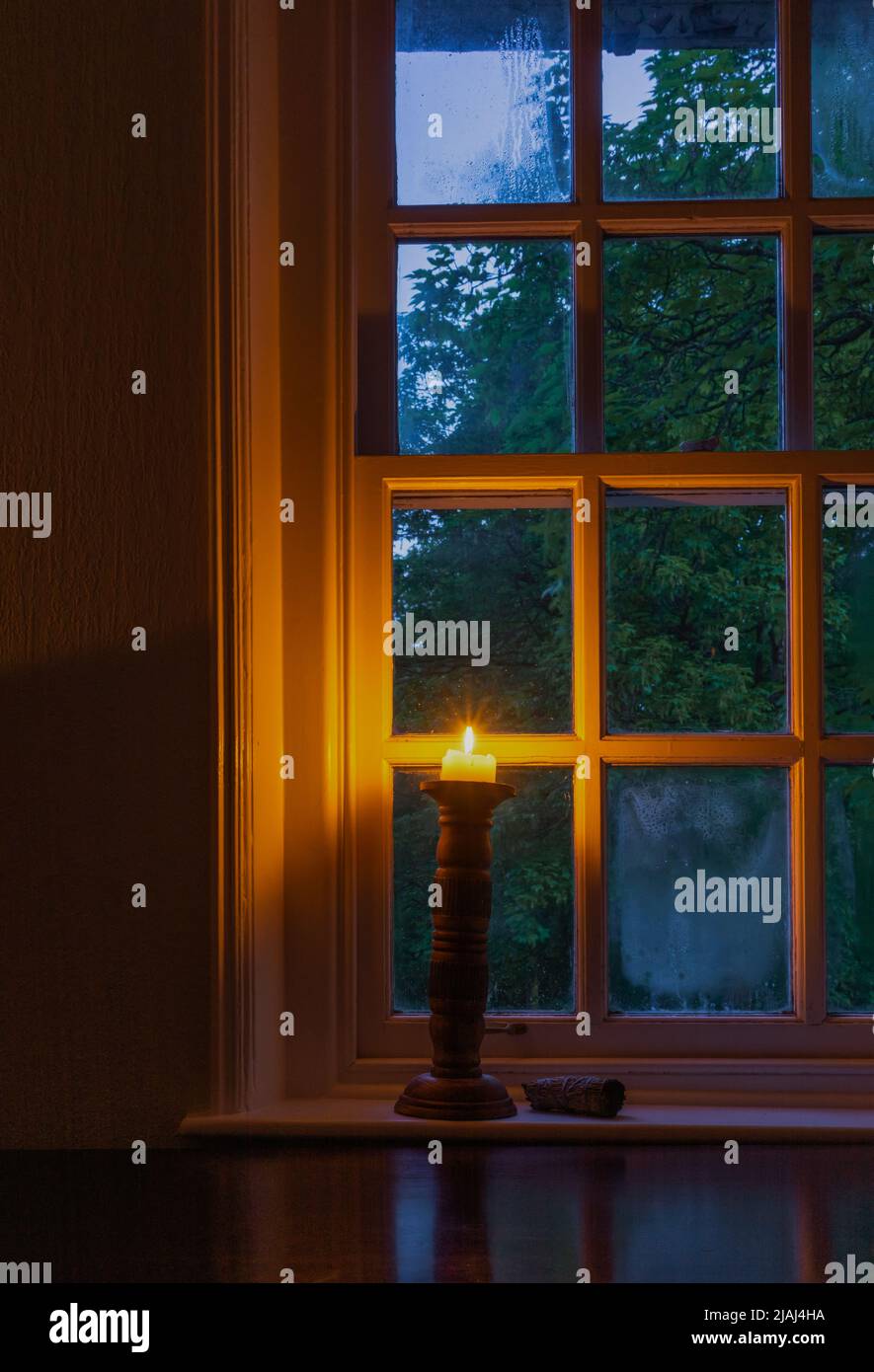 Una vela encendida en un portavelas de madera por una ventana durante la luz baja Foto de stock