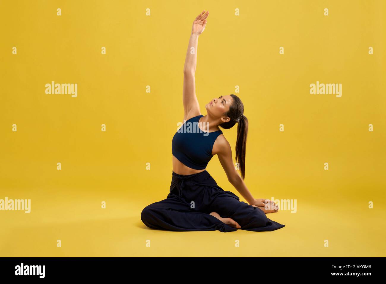 Vista frontal de una mujer flexible y bonita sentada en pose de yoga en el estudio. Chica delgada en negro activo posando con el brazo, estirado en el aire, aislado sobre fondo de estudio amarillo. Concepto de yoga. Foto de stock