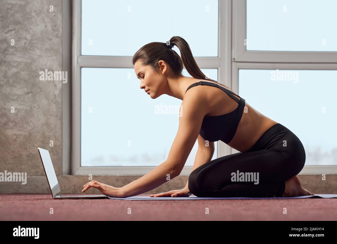 Vista lateral de la mujer en forma bonita haciendo yoga, mirando el  portátil en casa. Mujer atractiva en ropa deportiva sentada sobre las  rodillas, durante las clases en línea en casa con