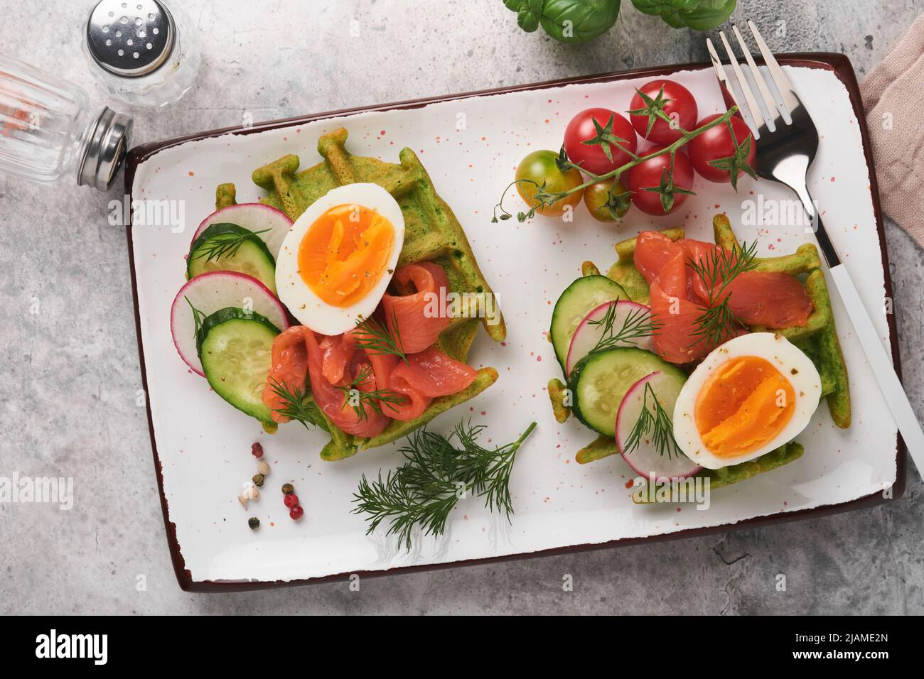 Gofres salados. Desayuno con gofres verdes, ajo o espinacas silvestres con huevo, tomate y salmón en el plato para el desayuno sobre fondo de mesa de hormigón gris. Ligh Foto de stock