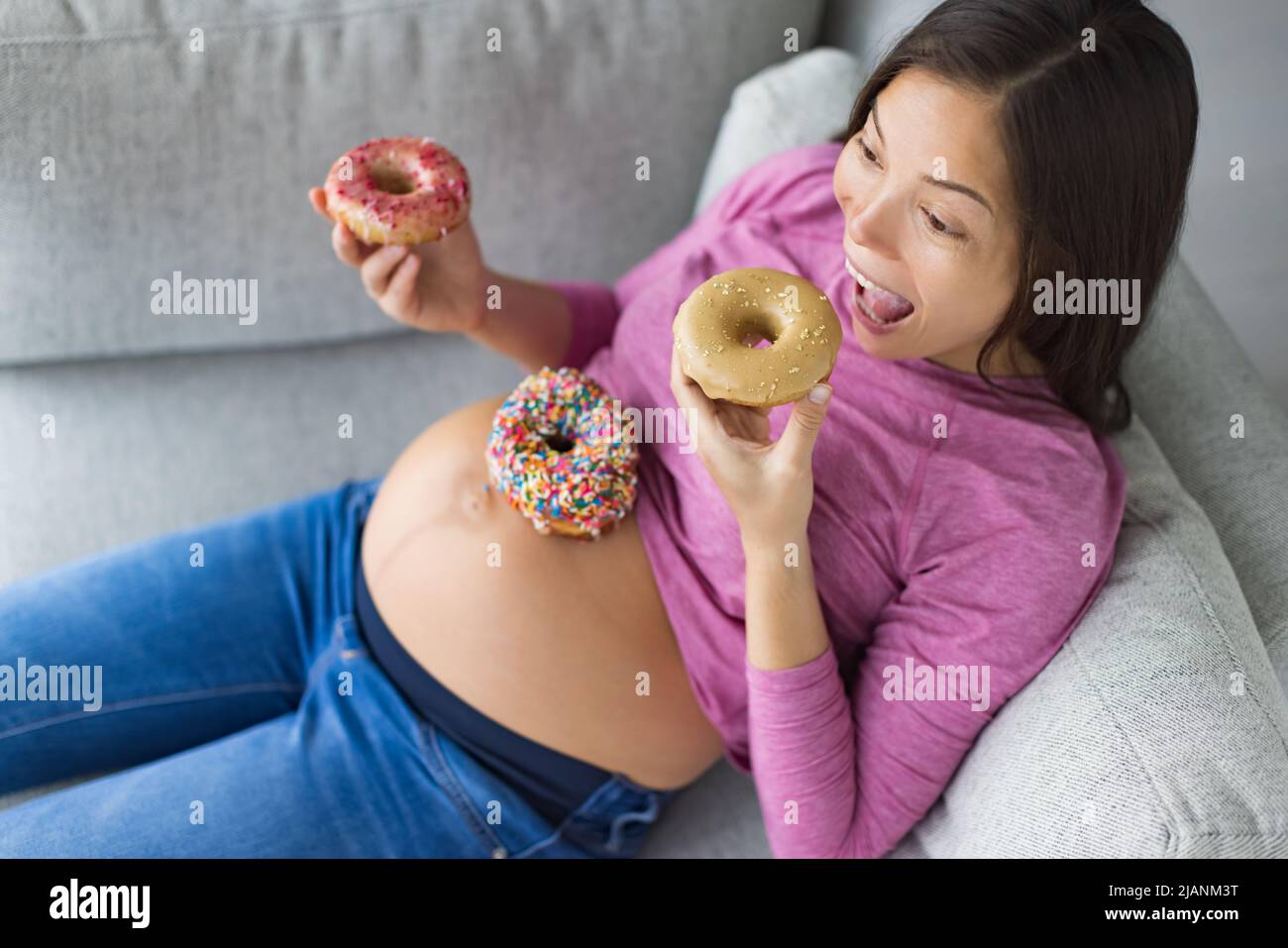 Embarazo alimentos antojos graciosa mujer asiática embarazada comer donuts en casa hambriento de pasteles de postre y aperitivos dulces. Diabetes gestacional, nutrición Foto de stock