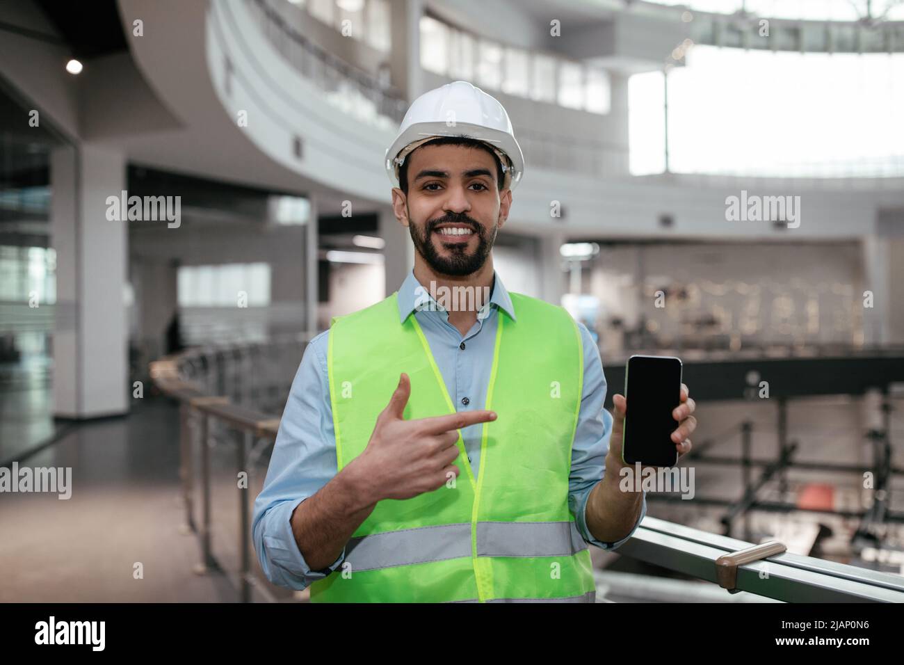 Alegre joven ingeniero islámico constructor con barba en la ropa protectora y el casco duro apunta con el dedo al smartphone Foto de stock