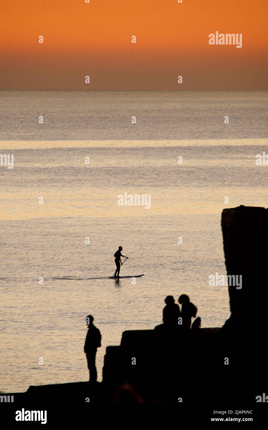 Siluetas formas de personas sentadas y de pie en un gryne en Brighton Beach, después de haber visto la puesta de sol. El cielo se aglow con la luz fadigna del día. Brighton, Inglaterra, Reino Unido Foto de stock