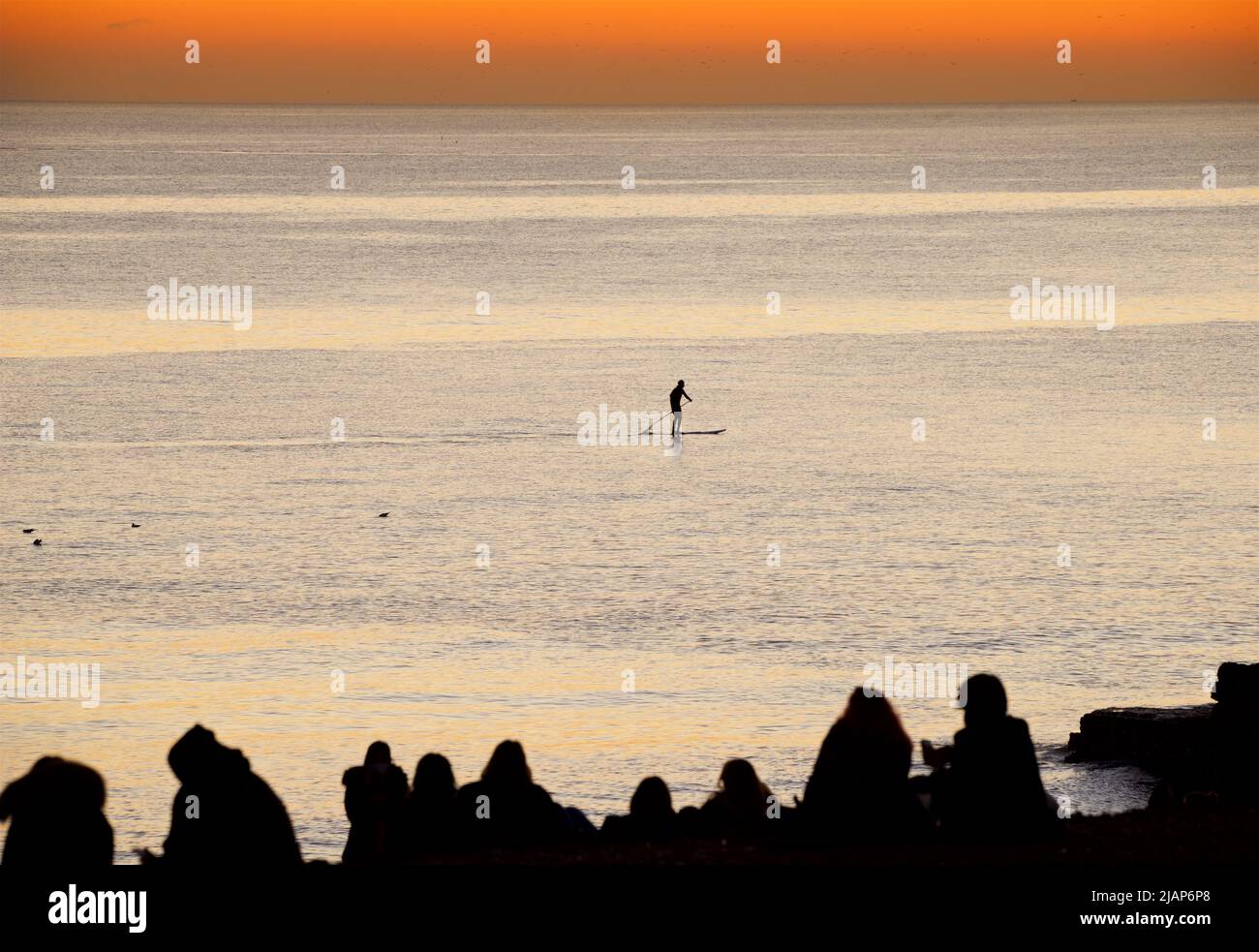 Siluetas formas de personas sentadas y de pie en un gryne en Brighton Beach, después de haber visto la puesta de sol. El cielo se aglow con la luz fadigna del día. Brighton, Inglaterra, Reino Unido Foto de stock
