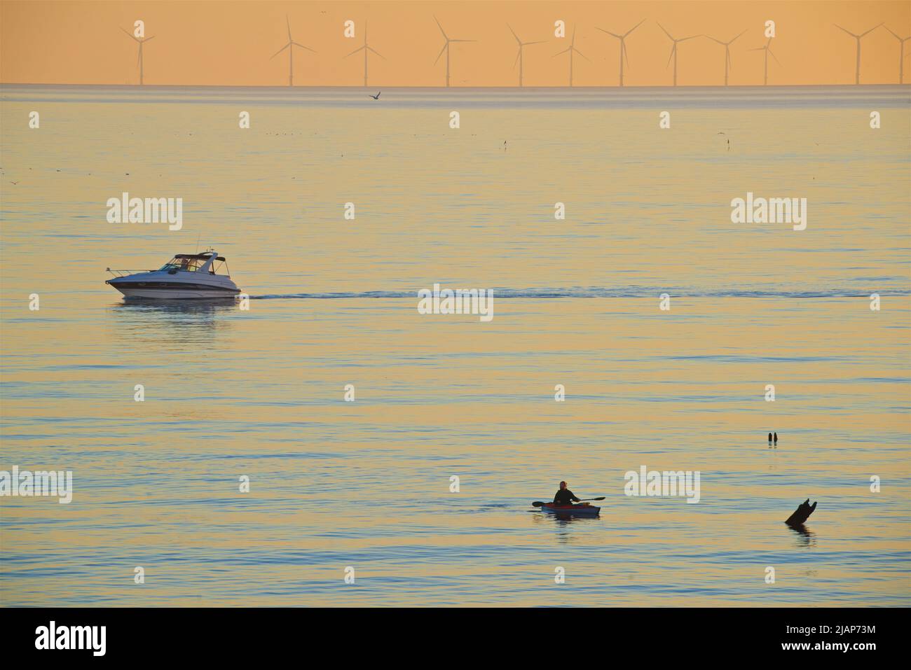 Silueta de un hombre en un kayak y un pequeño barco de motor en el mar. Aguas tranquilas, canal inglés. Brighton, Inglaterra, Reino Unido Las turbinas del Parque Eólico de Rampion en el horizonte lejano. Foto de stock