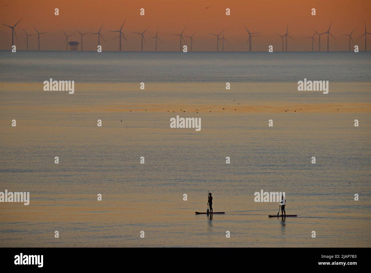 Pareja de surf de remo en el mar frente a Brighton, East Sussex, Inglaterra, con el parque eólico de Rampion en el horizonte lejano. Siluetas al atardecer. Foto de stock