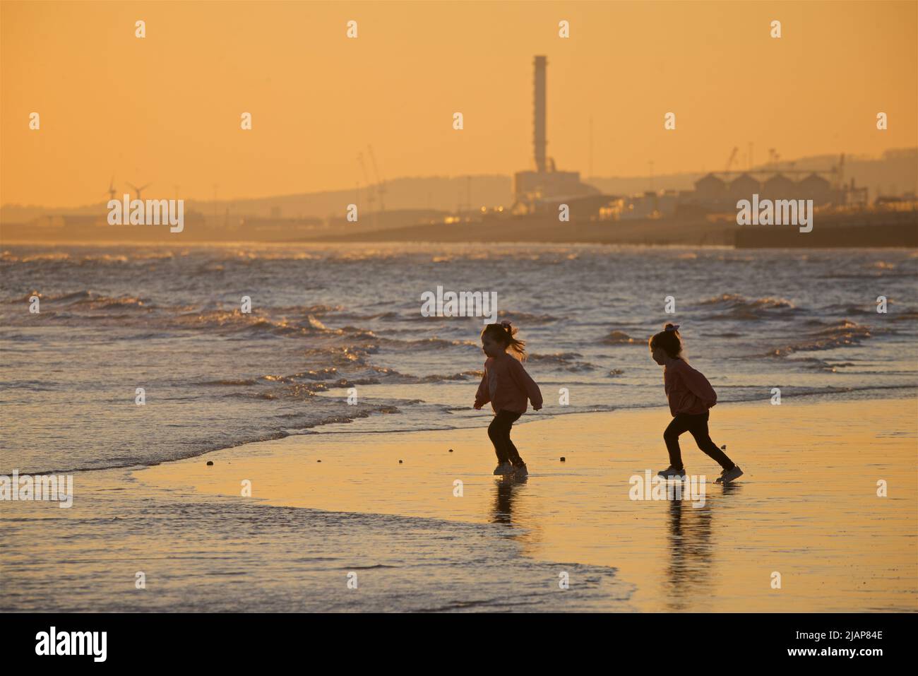 Dos niñas caminando a lo largo de la costa en marea baja, Brighton, Sussex, Inglaterra, Reino Unido. Puesta de sol / atardecer. La central eléctrica de Shoreham en segundo plano. Foto de stock