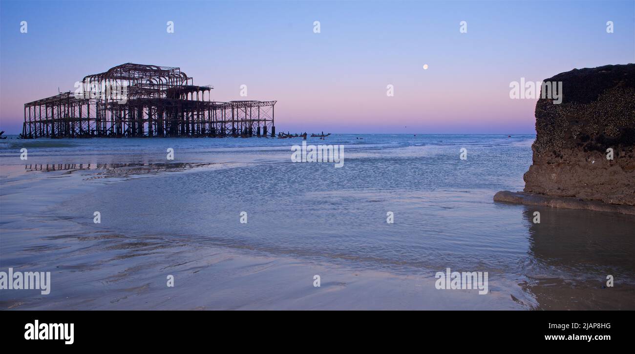 Restos oxidados del dilapidado West Pier en la marea baja del amanecer. Brighton & Hove, Sussex, Inglaterra, Reino Unido. Luna llena. Groyne y piscina de aguas poco profundas en el primer plano. Foto de stock