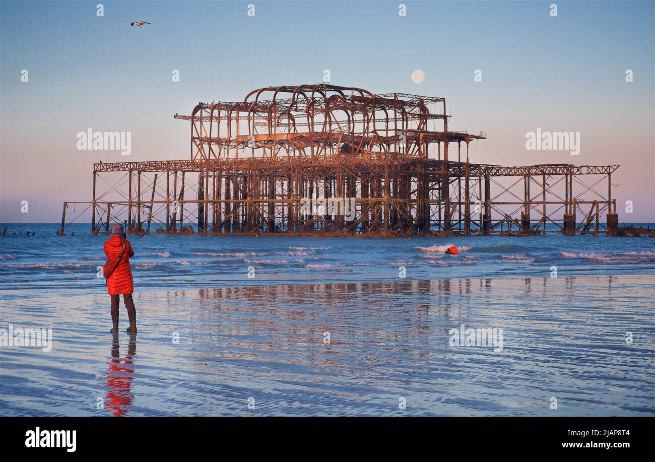Restos oxidados del dilapidado West Pier en la marea baja del amanecer. Brighton & Hove, Sussex, Inglaterra, Reino Unido. Luna llena. Mujer con abrigo rojo tomando fotografía del muelle. Foto de stock