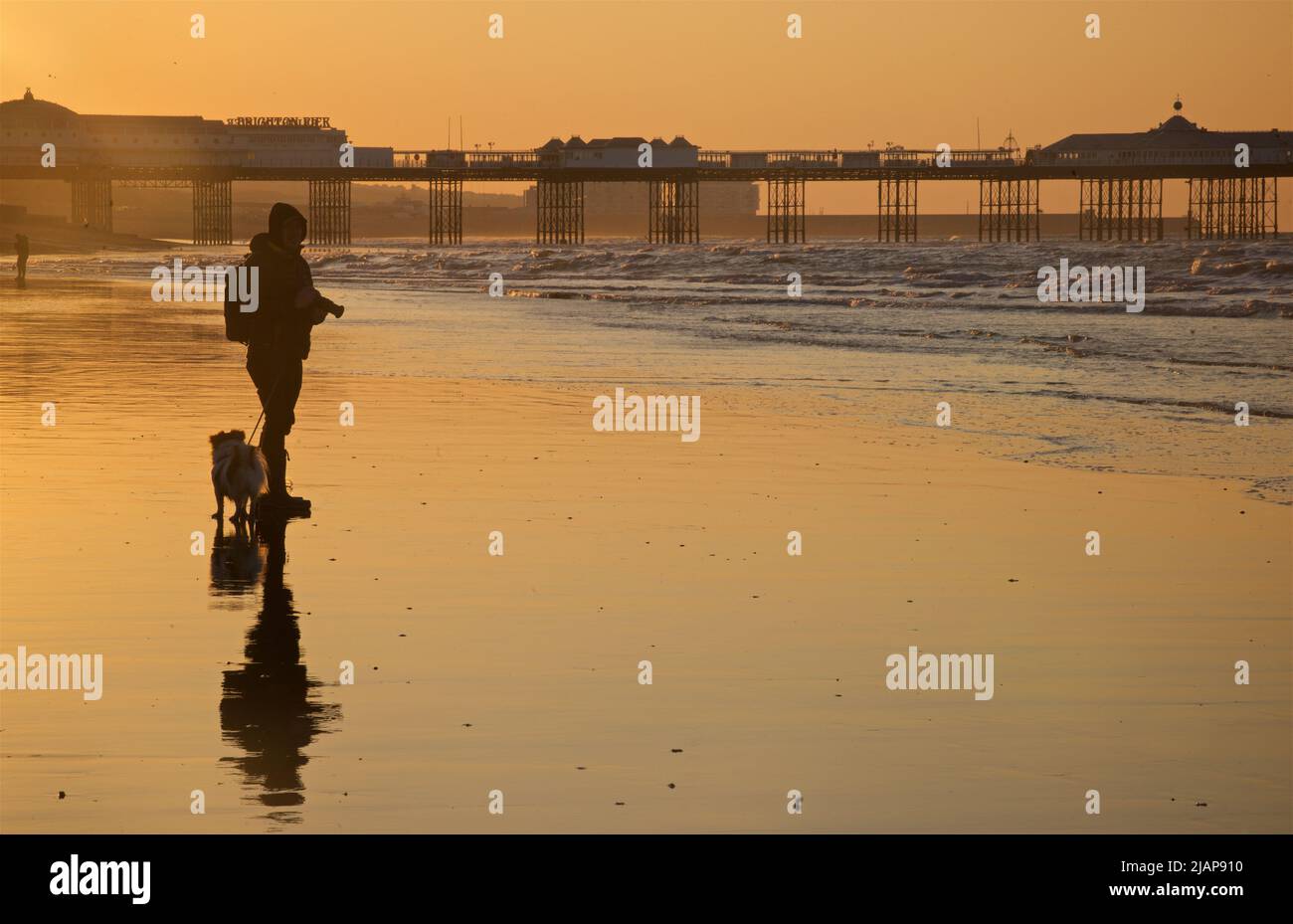 Siluetas del amanecer de personas en la playa en marea baja, Brighton & Hove, East Sussex, Inglaterra, Reino Unido. Paseo por la mañana con perro en la playa. Brighton Palace Pier en el fondo; fotógrafo con cámara. Foto de stock