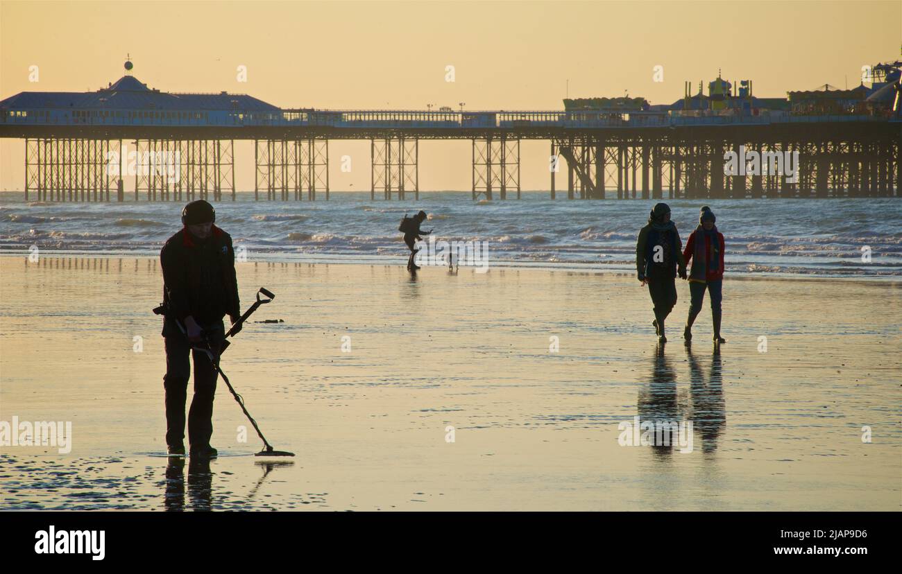 Detectorista de metales que se prospección en la arena en tiempo bajo. Playa al amanecer, Brighton & Hove, Sussex, Inglaterra, Reino Unido. Paseantes caminando por la arena, algunos con un perro; Palace Pier más allá. Foto de stock