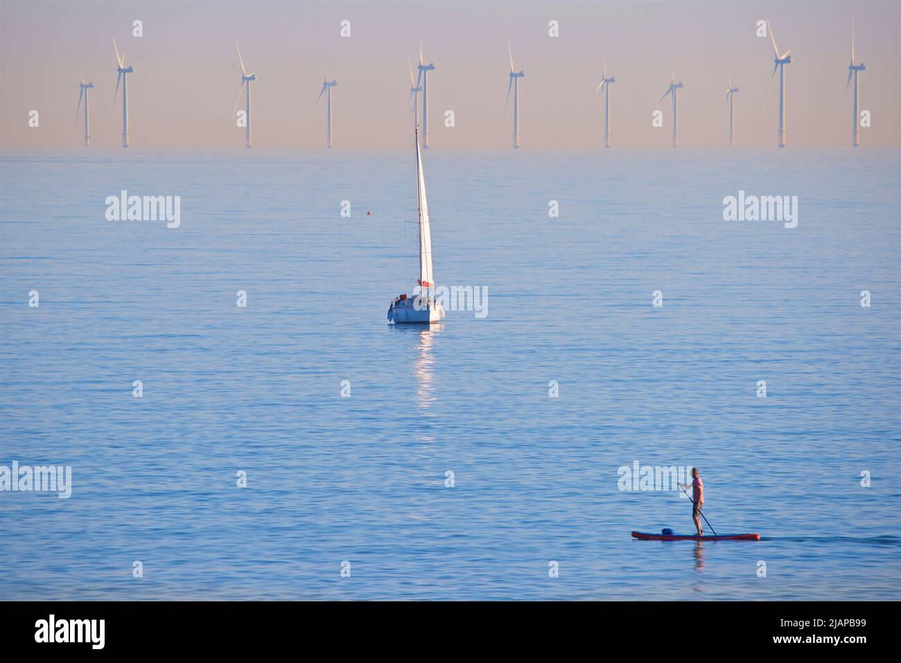 Turbinas del parque eólico marino Rampion ubicado en la costa de Sussex, Inglaterra, Reino Unido. Encargado y producciendo energía renovable en 2018, el parque eólico de 116 turbinas costó £1,3 construir. Yate en el suelo medio; paddleboarder en primer plano. Foto de stock