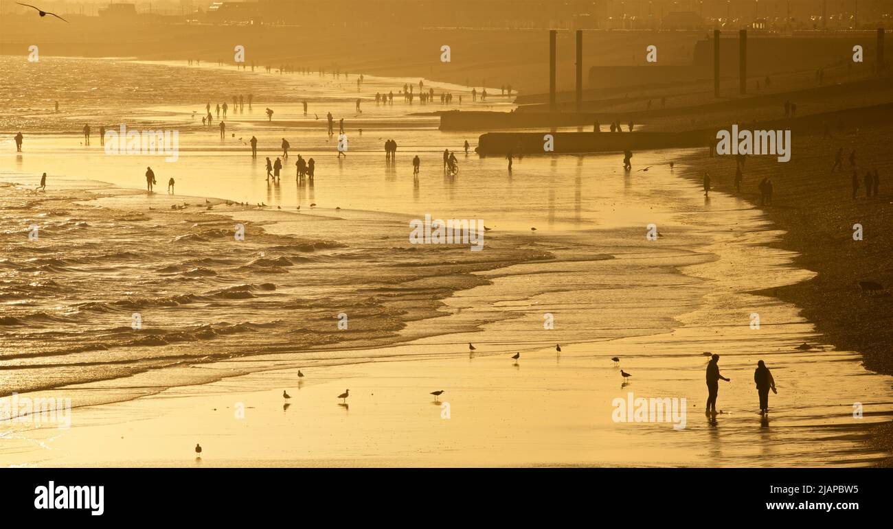 Siluetas de una pareja en primer plano, y otras personas en la playa en marea baja, Brighton & Hove, East Sussex, Inglaterra, Reino Unido. Los pilares verticales más allá a la derecha representan los restos del viejo West Pier. Foto de stock