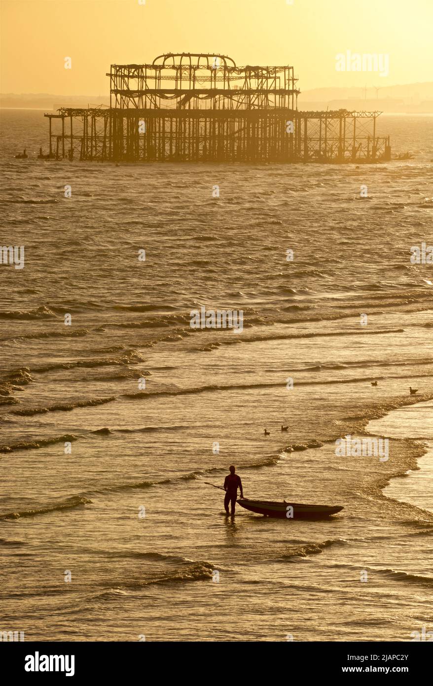 Siluetas de hombre con kayak en la costa en marea baja, Brighton & Hove, East Sussex, Inglaterra, Reino Unido. Restos del dilapidado West Pier más allá. Foto de stock