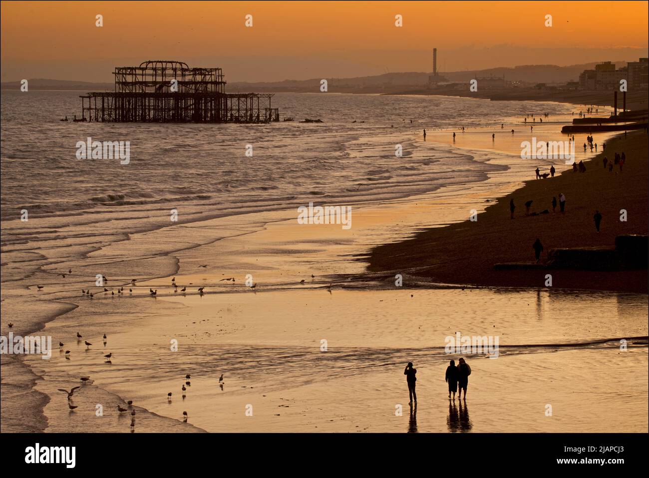 Siluetas de amigos juntos y otras personas en la playa en marea baja, Brighton & Hove, East Sussex, Inglaterra, Reino Unido. Fotografiado desde el muelle del Palacio. Shoreham Power Station en la distancia. Foto de stock