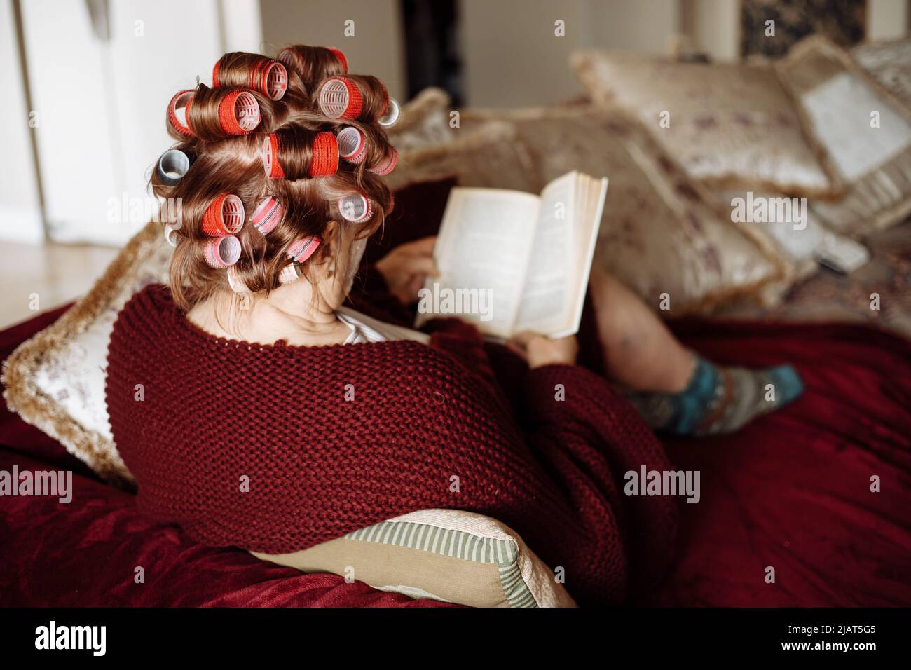 Vista posterior de la anciana aplicando coloridos cabellos-rizadores, llevando un cárdigan vinoso, sentado en el sofá, libro de lectura. Foto de stock
