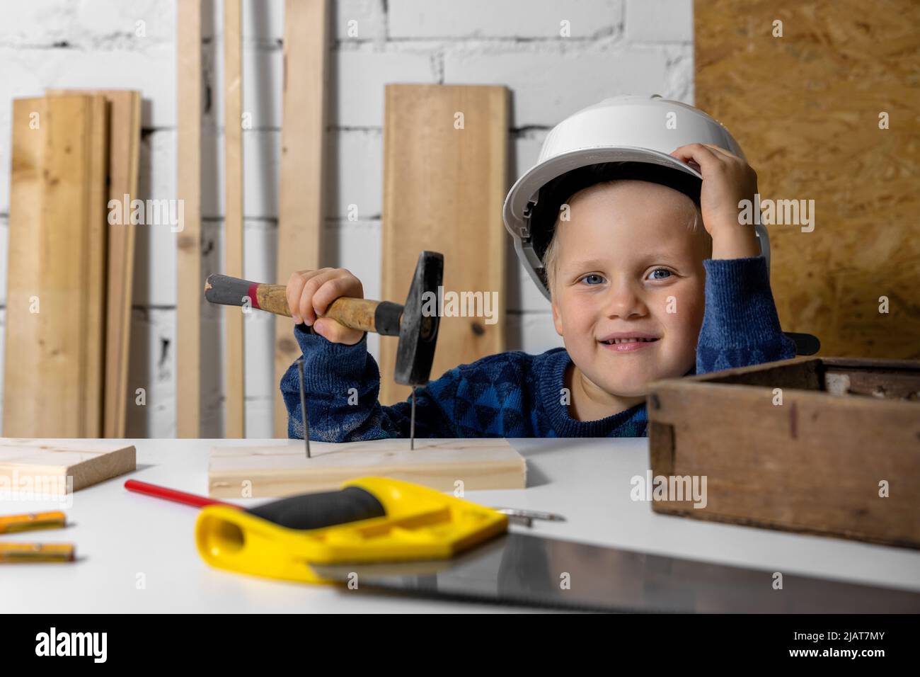 feliz niño sonriente con casco y martillo en la mano en el taller de carpintería Foto de stock
