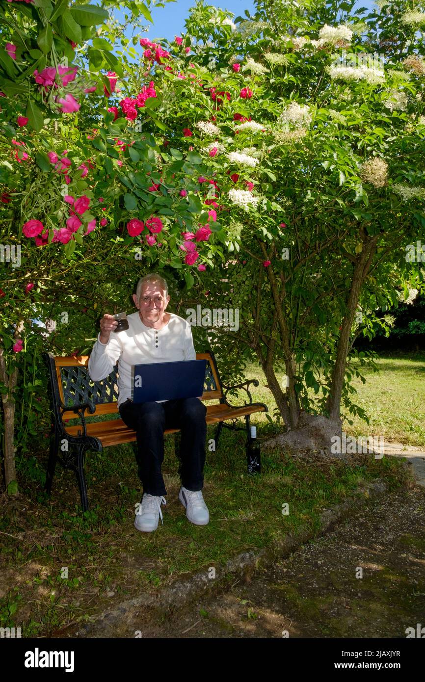 hombre maduro sentado en el jardín con ordenador portátil que trabaja desde casa bajo la sombra de un cenador natural de rosas sosteniendo una copa de vino Foto de stock