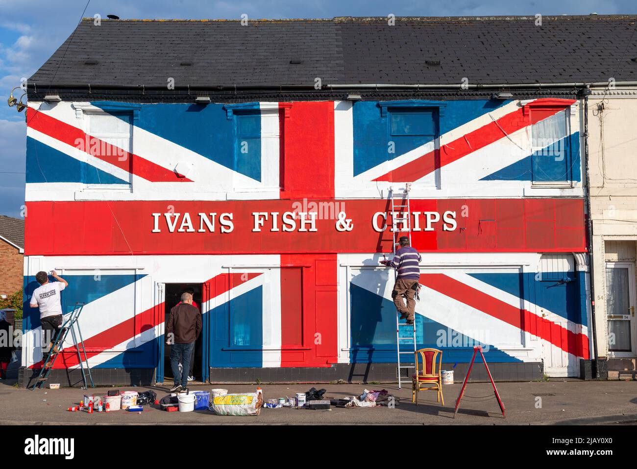 Cradley Heath, West Midlands, Reino Unido. 1st de junio de 2022. Warren Rudge, propietario de la tienda de chips, pone los toques finales en la bandera de la Unión que él y su hijo James han pintado en su tienda de pescado y fichas en Cradley Heath, West Midlands. La bandera pintada cubre toda la fachada de Ivan's Fish and Chips incluyendo las ventanas, y ha tomado todo el día para completarse. 'Ivan's Fish and Chips ha estado sirviendo pescado y patatas fritas durante 65 años - no tan largo como Su Majestad sin embargo. Pero he oído que la Reina es parcial al pescado y a las patatas fritas.' dice Warren. Crédito: Peter Lodeman/Alamy Live News Foto de stock