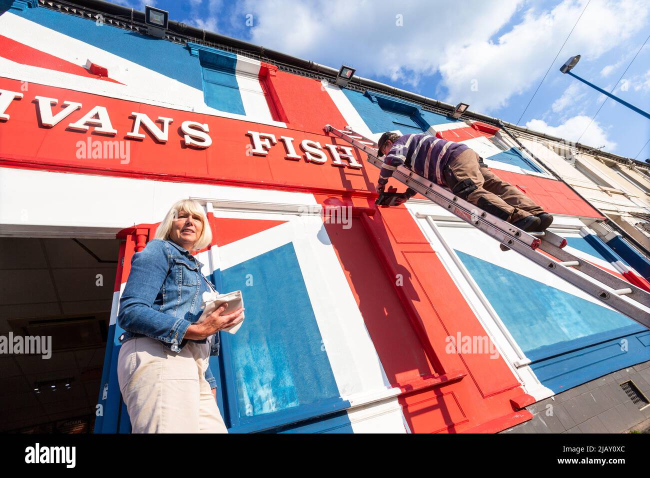 Cradley Heath, West Midlands, Reino Unido. 1st de junio de 2022. Warren Rudge, propietario de la tienda de chips, pone los toques finales en la bandera de la Unión que él y su hijo James han pintado en su tienda de pescado y fichas en Cradley Heath, West Midlands. La bandera pintada cubre toda la fachada de Ivan's Fish and Chips incluyendo las ventanas, y ha tomado todo el día para completarse. 'Ivan's Fish and Chips ha estado sirviendo pescado y patatas fritas durante 65 años - no tan largo como Su Majestad sin embargo. Pero he oído que la Reina es parcial al pescado y a las patatas fritas.' dice Warren. Crédito: Peter Lodeman/Alamy Live News Foto de stock