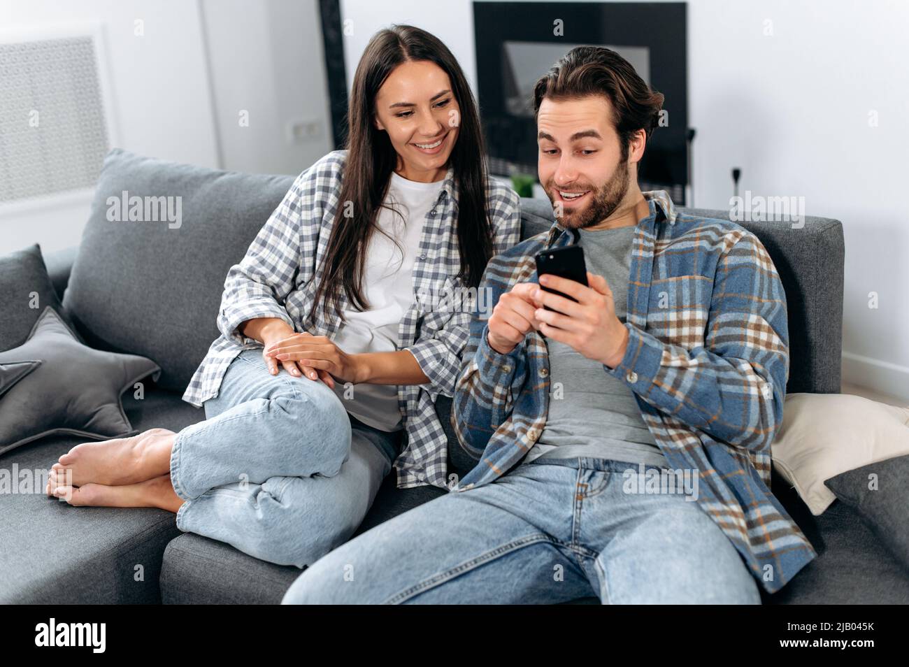 Feliz hombre y mujer enamorada vestida de ropa informal de moda, pasar  tiempo libre en casa juntos en el sofá, utilizar el teléfono, ver las  noticias, navegar por redes sociales, poner apuestas,