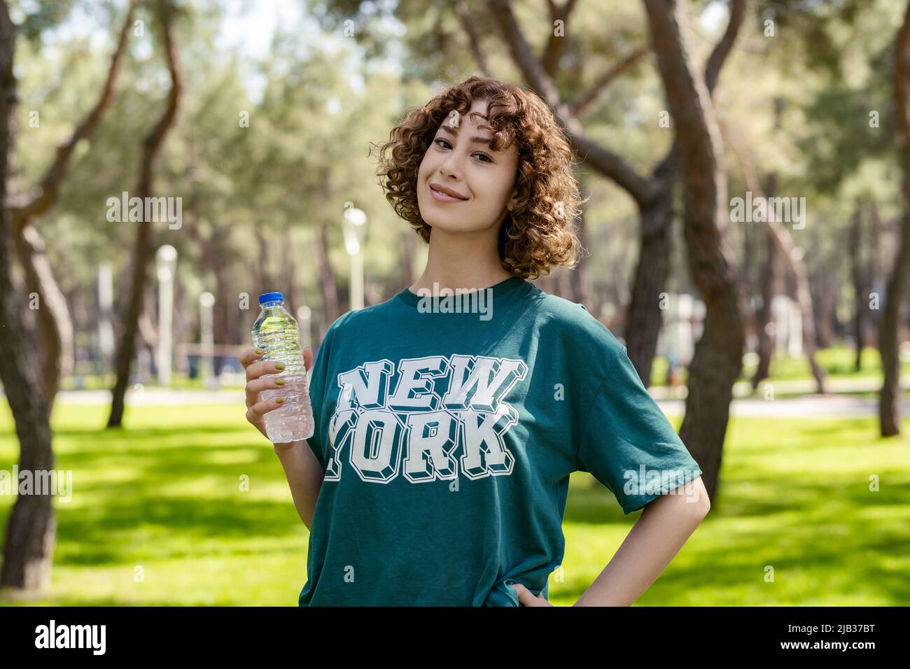 Hermosa mujer joven pelirroja con camiseta verde de pie en el parque de la ciudad, al aire libre sosteniendo una botella agua. Mirando la cámara. Deportes al aire libre, saludable l
