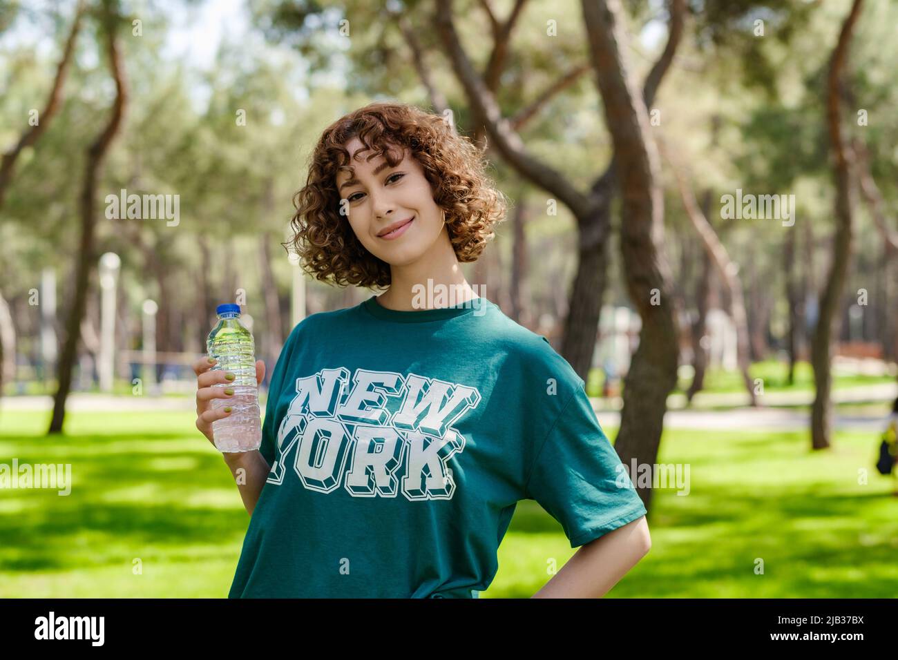 Feliz joven pelirroja con camiseta de pie en el parque de la ciudad, al aire sosteniendo una botella de agua. Mujer de fitness tomando un descanso después ejercicio