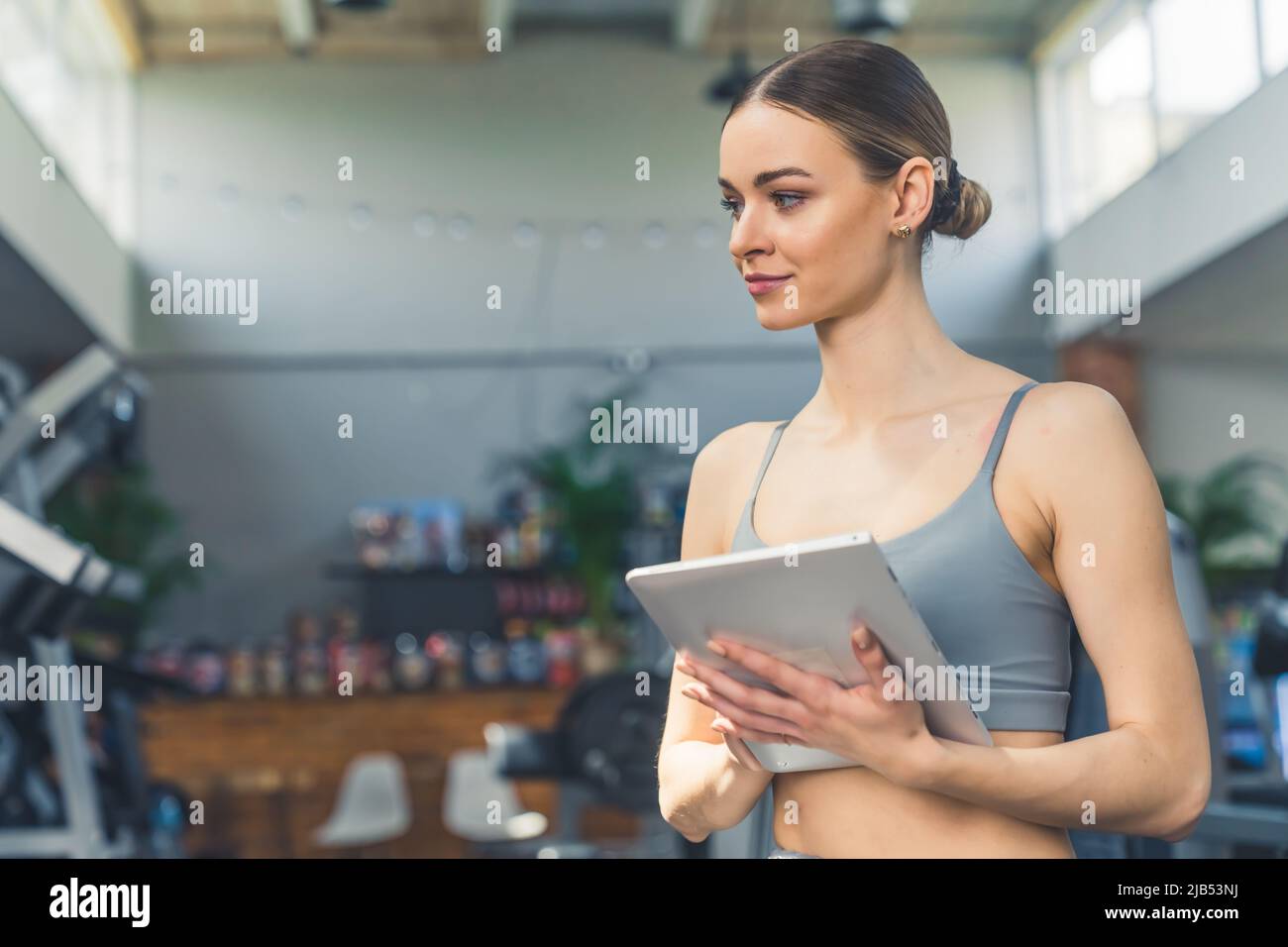 Retrato interior de una joven mujer blanca milenaria con el pelo de espalda y un sujetador deportivo gris con una tableta, creando un programa de entrenamiento para sus clientes. Fotografías de alta calidad Foto de stock