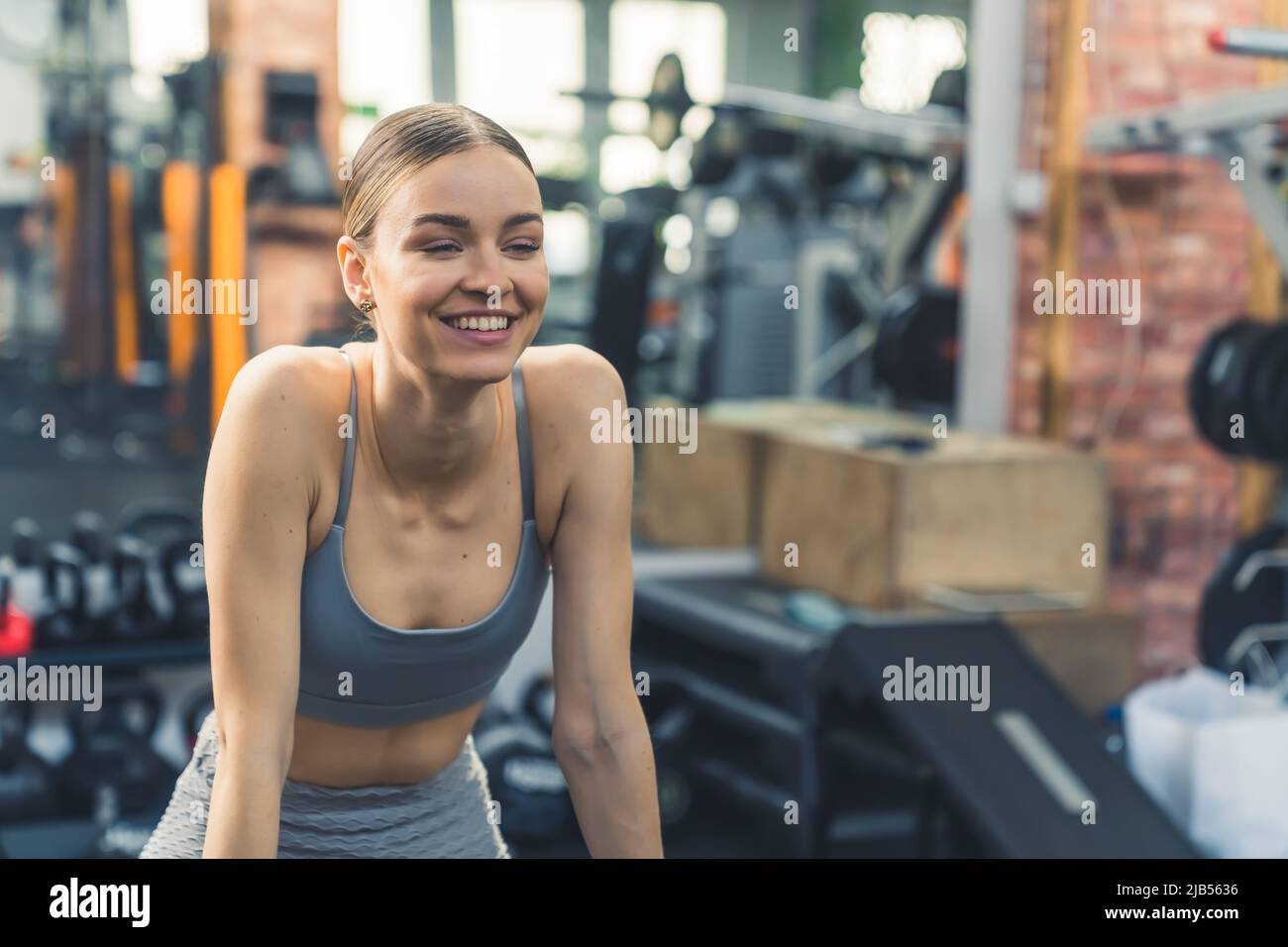 Tiro interior mediano en un gimnasio. Alegre mujer europea en su 20s en un sujetador deportivo y leggings mirando hacia delante y sonriendo después de un entrenamiento. Fotografías de alta calidad Foto de stock