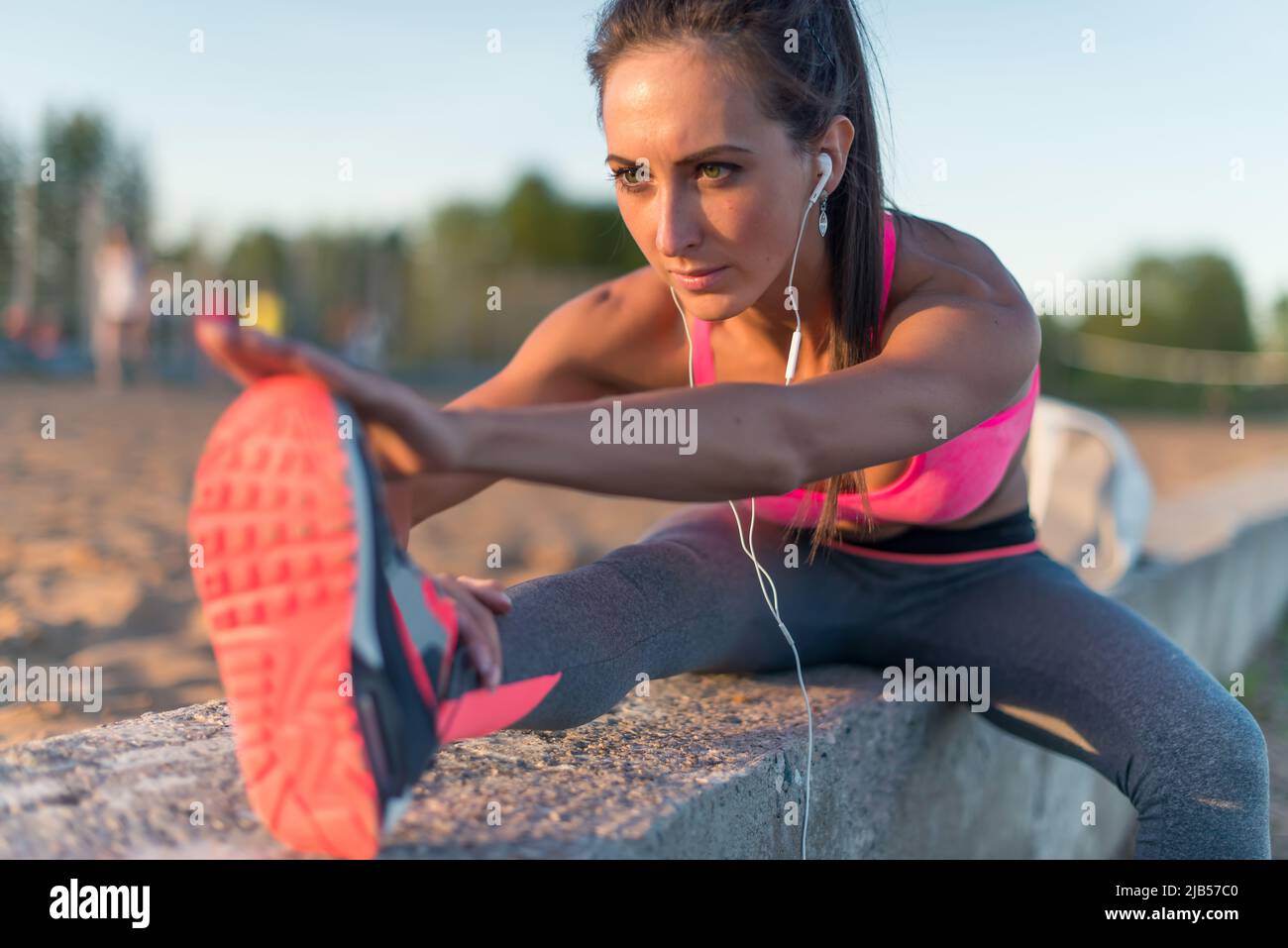 Modelo de fitness atleta chica de calentamiento estirando sus cuerdas,  pierna y espalda. Mujer joven haciendo ejercicio con auriculares escuchando  música al aire libre en la playa o Fotografía de stock -