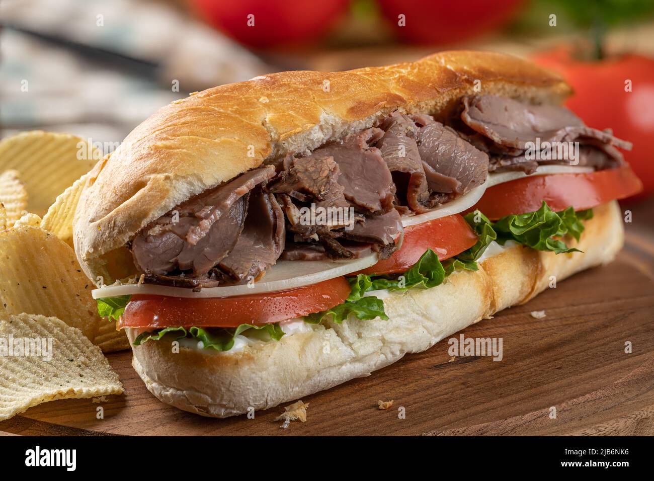 Cierre de un sándwich submarino con carne asada, queso, tomate y lechuga en un rollo de hoagie con patatas fritas en una tabla de cortar Foto de stock