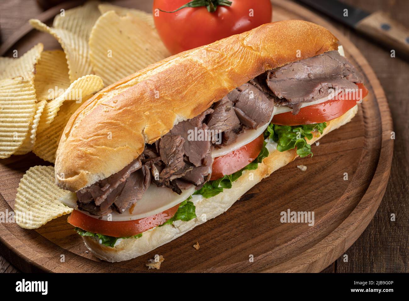Sándwich submarino con carne asada, queso, tomate y lechuga en un rollo de hoagie con patatas fritas en una tabla de cortar Foto de stock