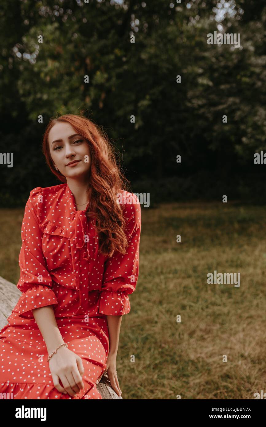 Retrato de mujer joven pelirroja en vestido corto de verano rojo con motas blancas sentado en la viga de madera de árbol caído seco Foto de stock