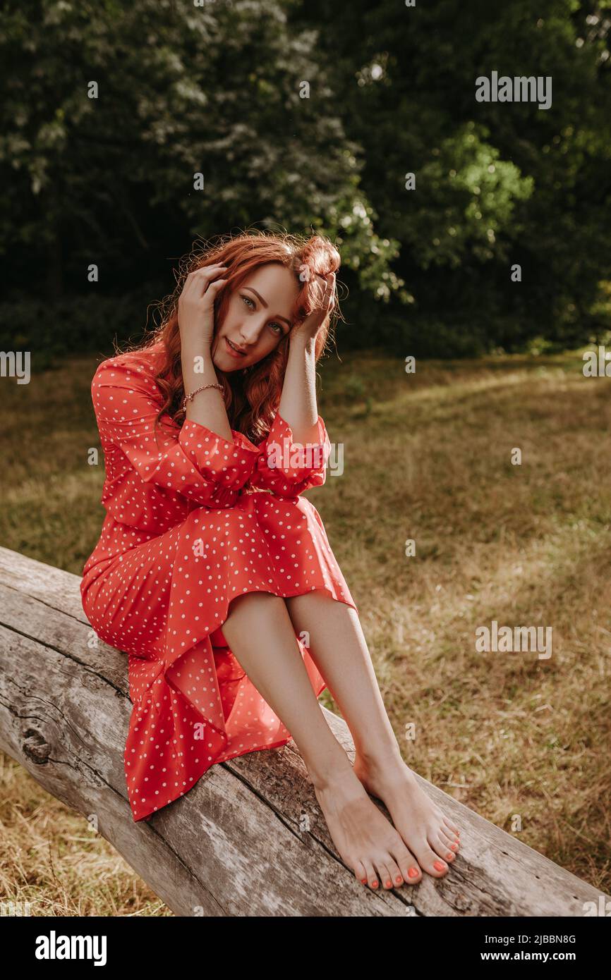 Una mujer adulta joven y bonita con vestido rojo con puntos blancos y pelo rojo sentado en un árbol caído seco y posando en una cámara. Modelo femenino tocando su h