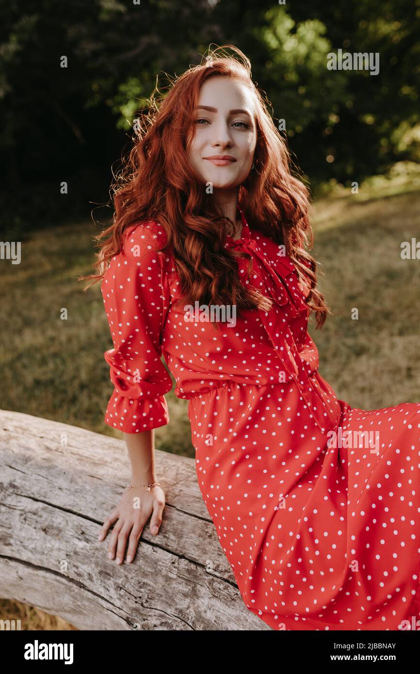 prima Alicia pausa Una mujer adulta joven y bonita con un vestido rojo con puntos blancos y  pelo rojo sentado en un árbol caído seco y posando en una cámara. Modelo  femenino tocando su h
