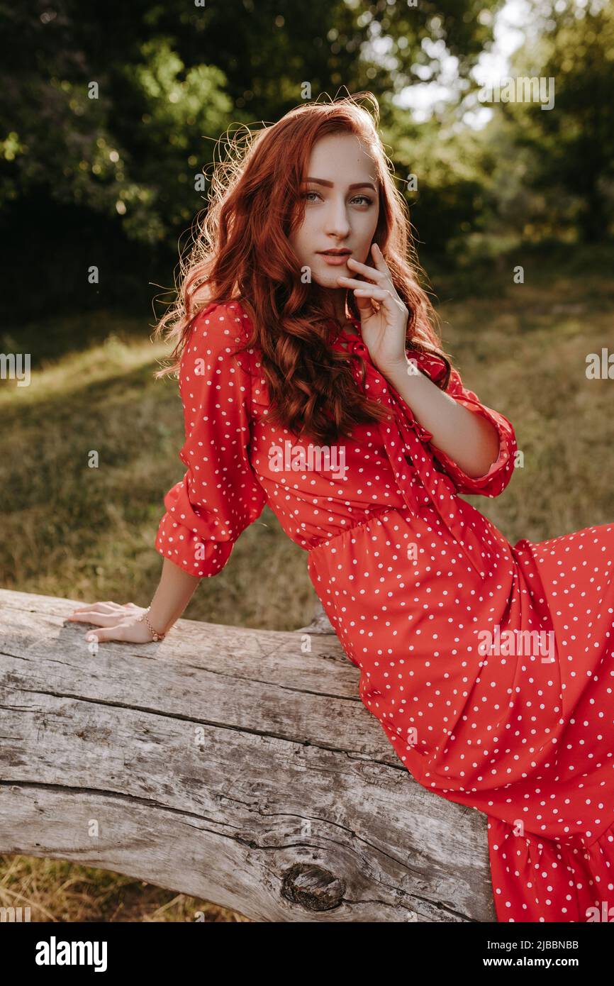 prima Alicia pausa Una mujer adulta joven y bonita con un vestido rojo con puntos blancos y  pelo rojo sentado en un árbol caído seco y posando en una cámara. Modelo  femenino tocando su h
