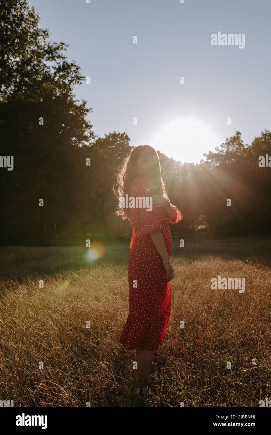 Mujer de vestido rojo de pie en el campo sobre el fondo del bosque. Mujer posando en una cámara a la luz del sol al aire libre Foto de stock