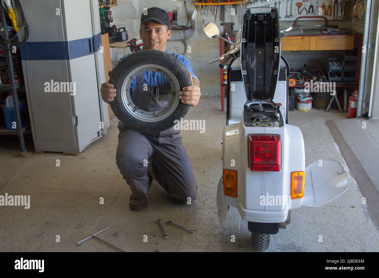 Imagen de un mecánico sonriente en su taller sosteniendo una rueda de la motocicleta que está reparando. Foto de stock
