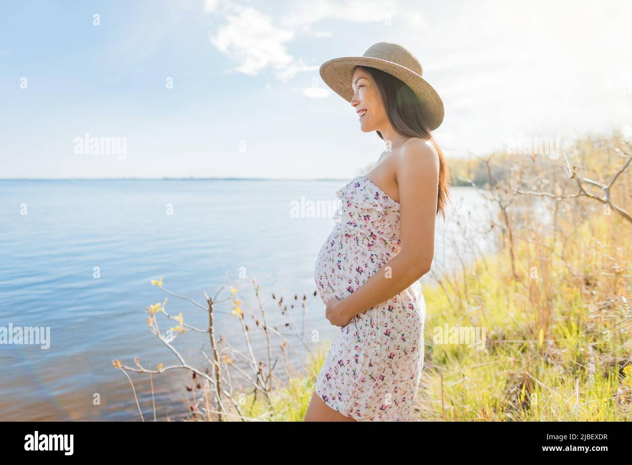 Mujer asiática embarazada que lleva sombrero durante la primavera de la naturaleza caminar por el lago relajarse disfrutando de la vista del paisaje de agua natural. Feliz embarazo al aire libre Foto de stock