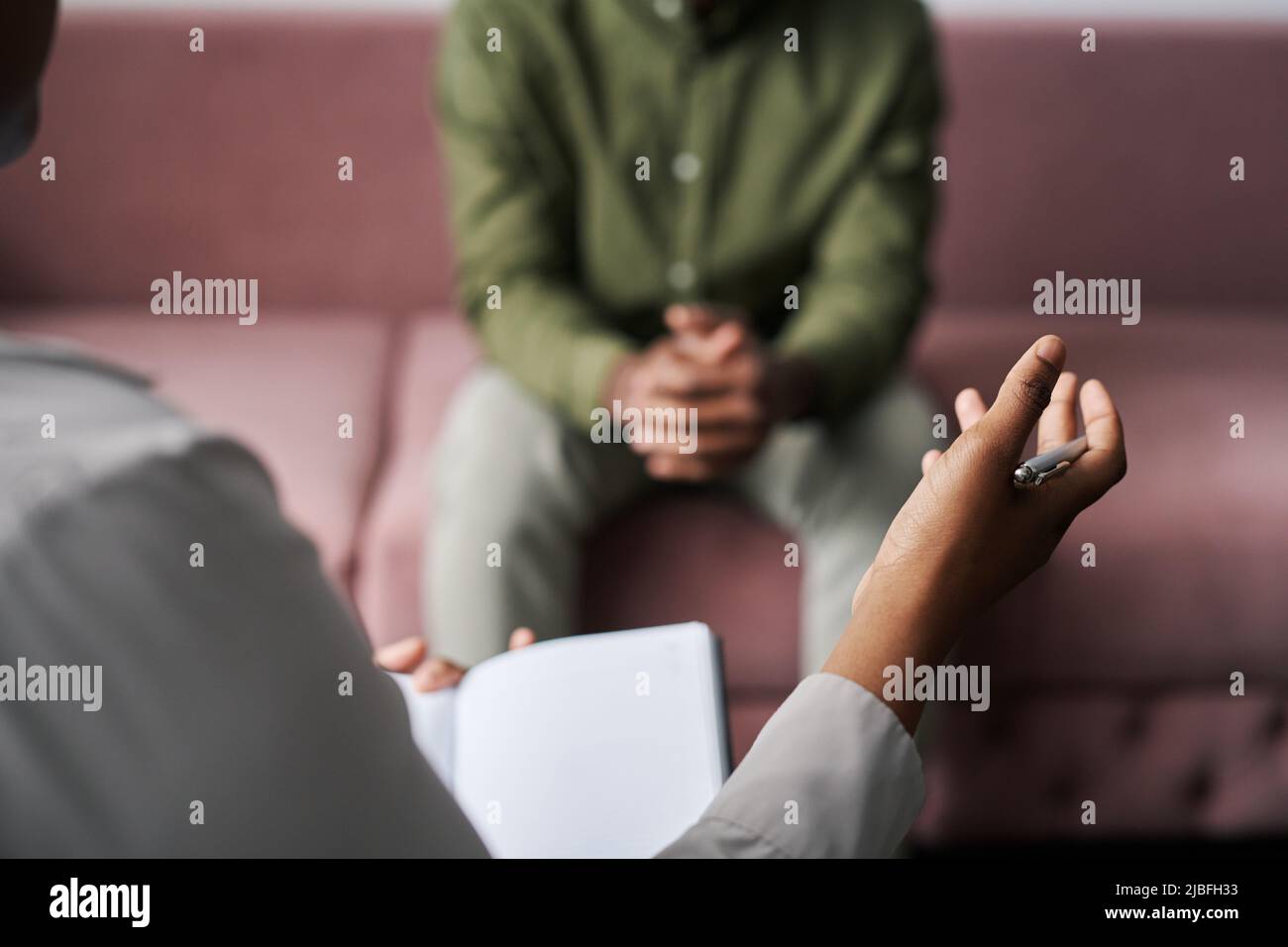 Mano del joven psicoanalista afroamericano con pluma que da consejos a un paciente masculino sentado en el sofá frente a ella durante la sesión Foto de stock