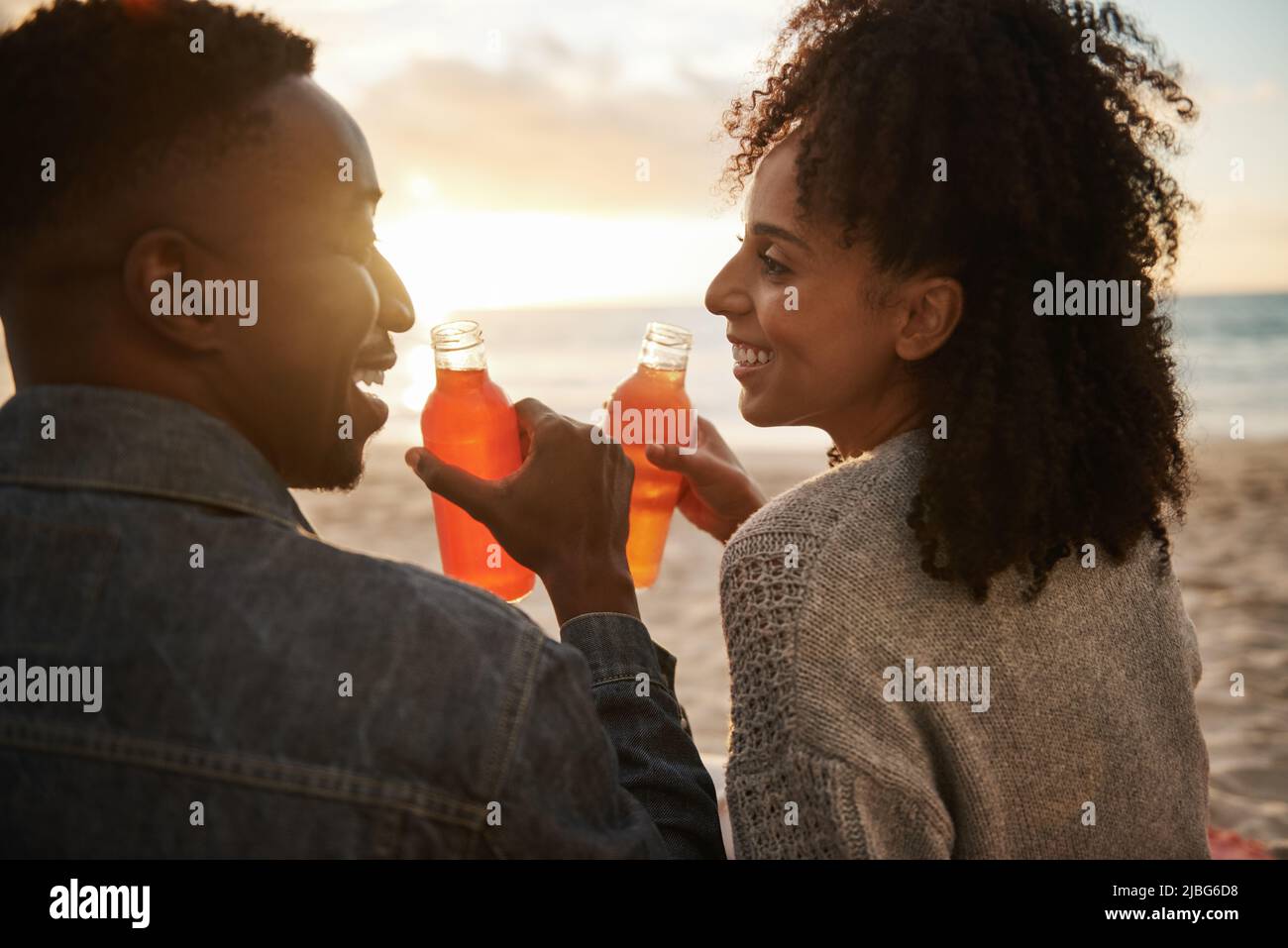 Sonriendo joven pareja multiétnica bebiendo jugo en una playa al atardecer Foto de stock