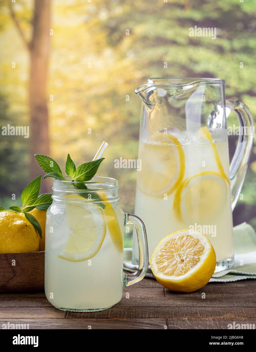 Limonada con rodajas de limón y menta en un vaso con una jarra y limonnas en el bacground sobre una mesa de madera con fondo rural Foto de stock