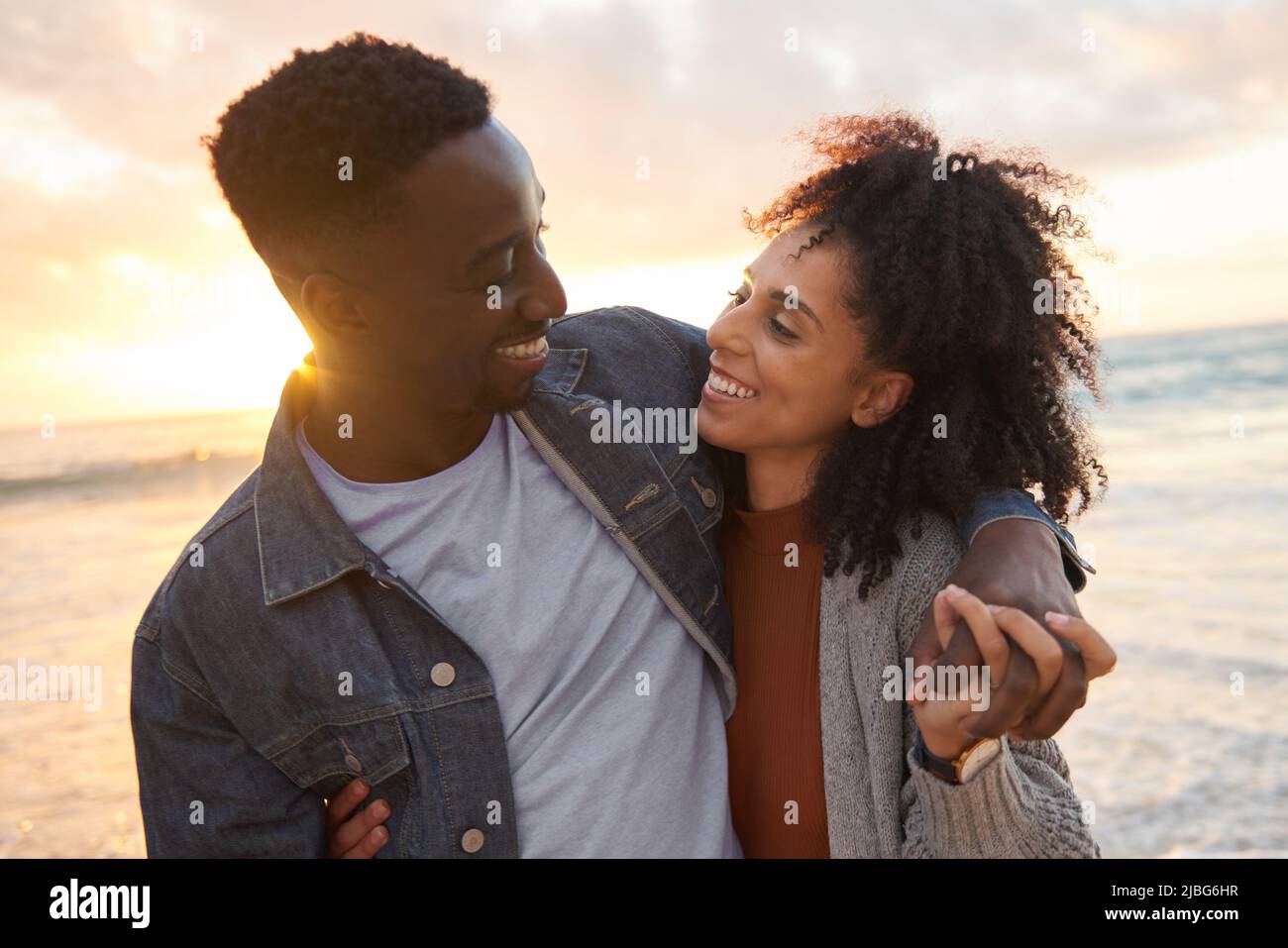 Sonriente pareja multiétnica de pie en el brazo en una playa al atardecer Foto de stock