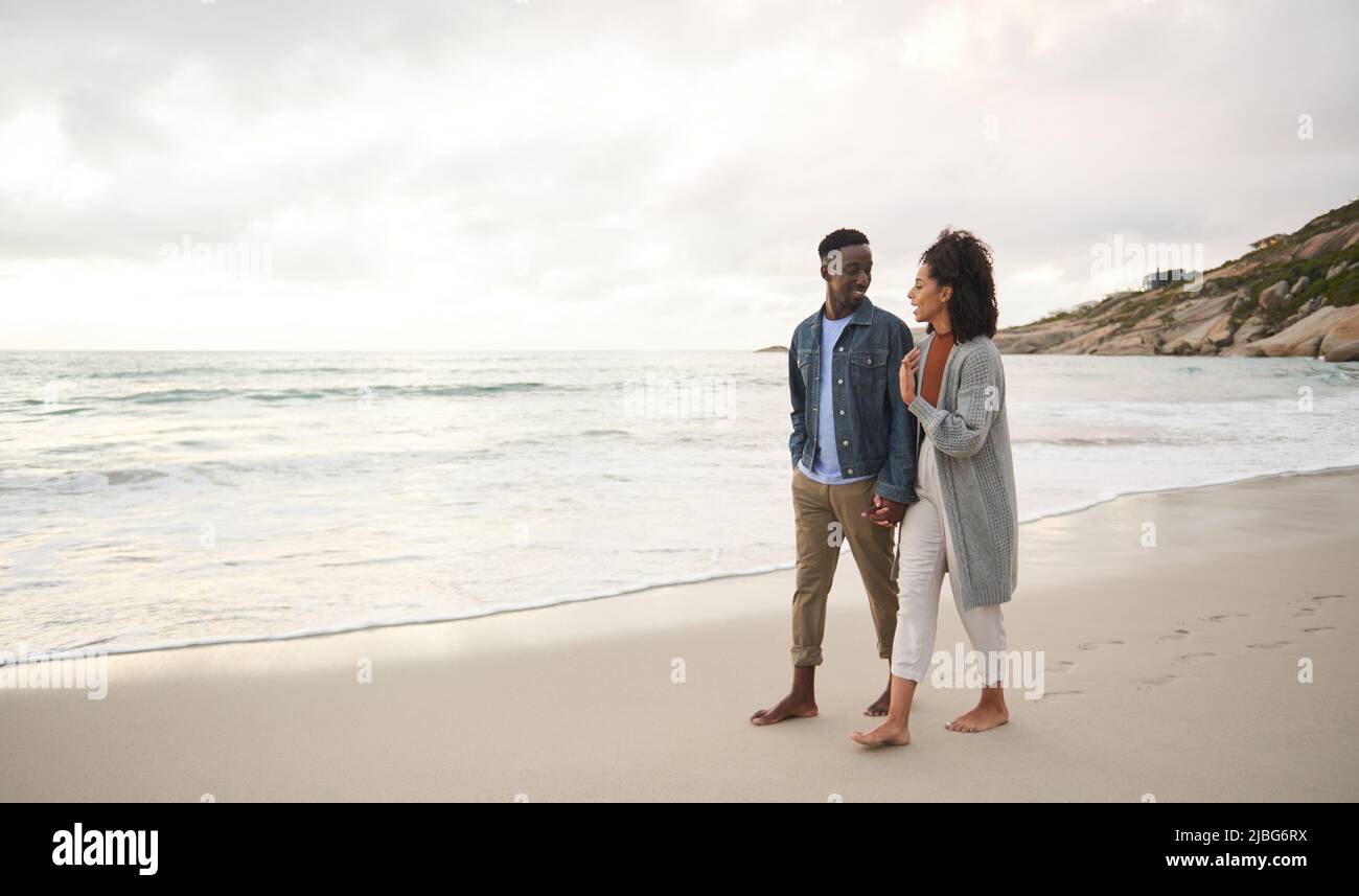 Sonriendo joven pareja multiétnica sosteniendo las manos mientras camina en una playa de arena Foto de stock