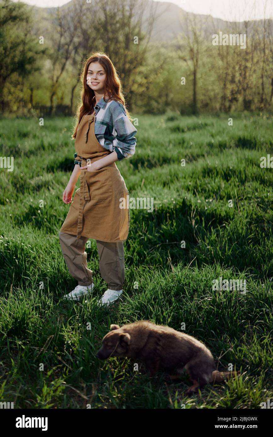 Una mujer campesina en su ropa de trabajo, camisa de cuadros y delantal, se  encuentra en el campo en el césped verde y sonríe en la puesta de sol  después de un
